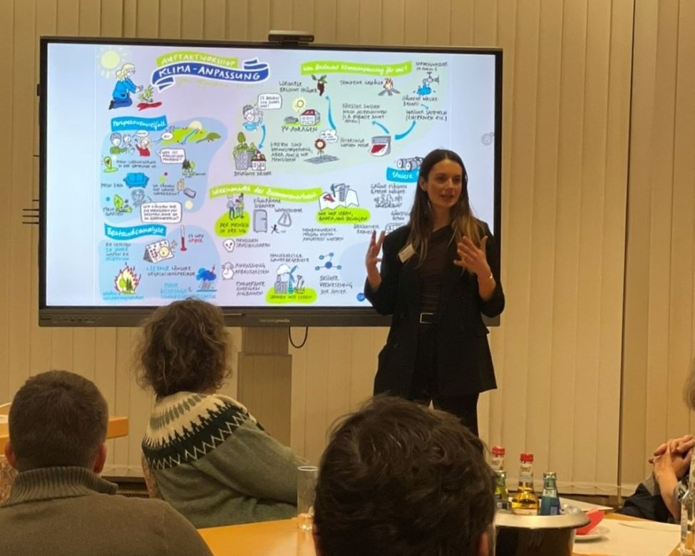 Zu sehen ist ein Bild des 1. Workshop. Eine Moderatorin erläutert ein Schaubild auf einem Digitalen Display.